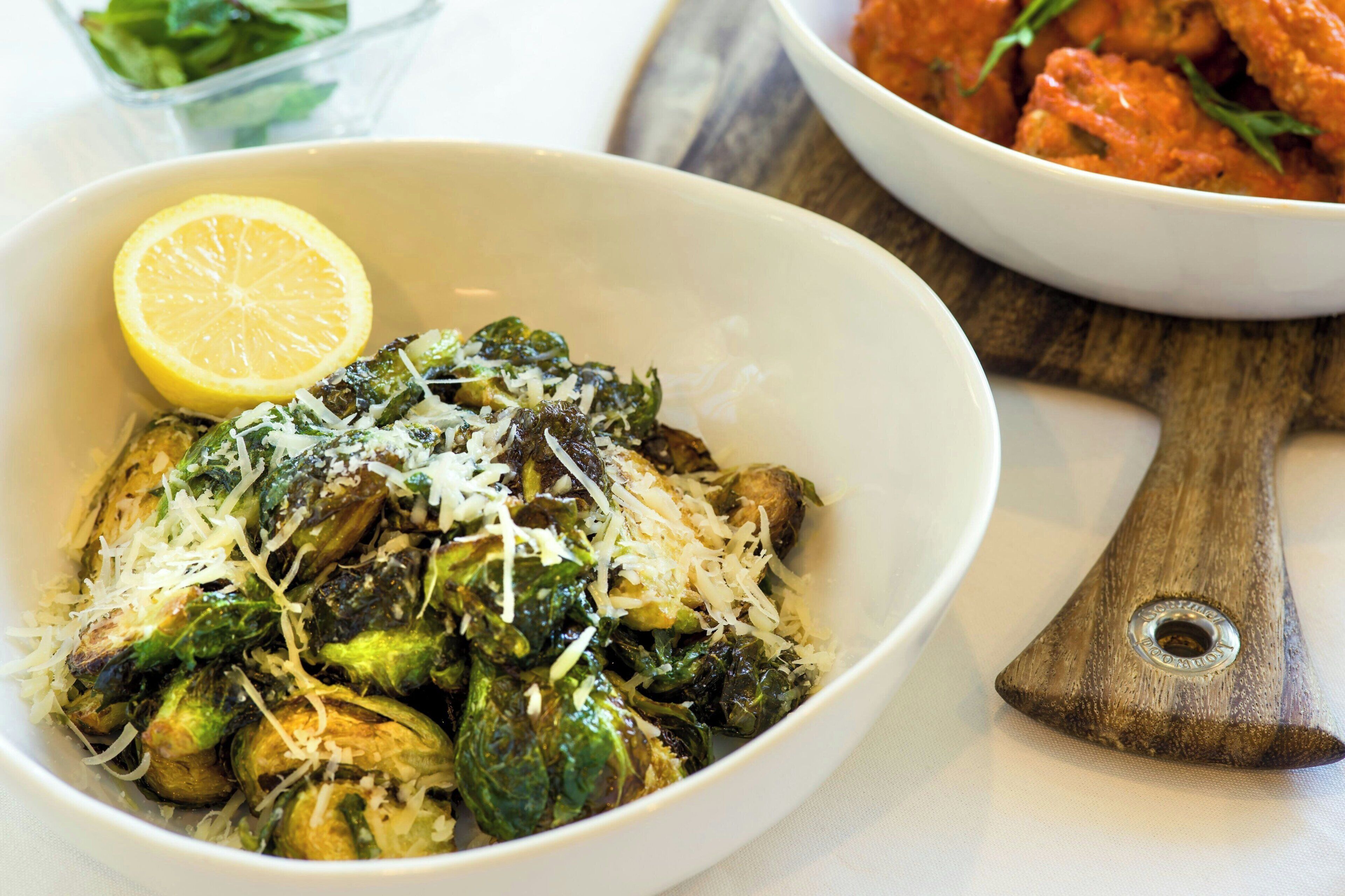 A bowl of cooked Brussels sprouts with cheese and a lemon wedge, on a wooden cutting board.