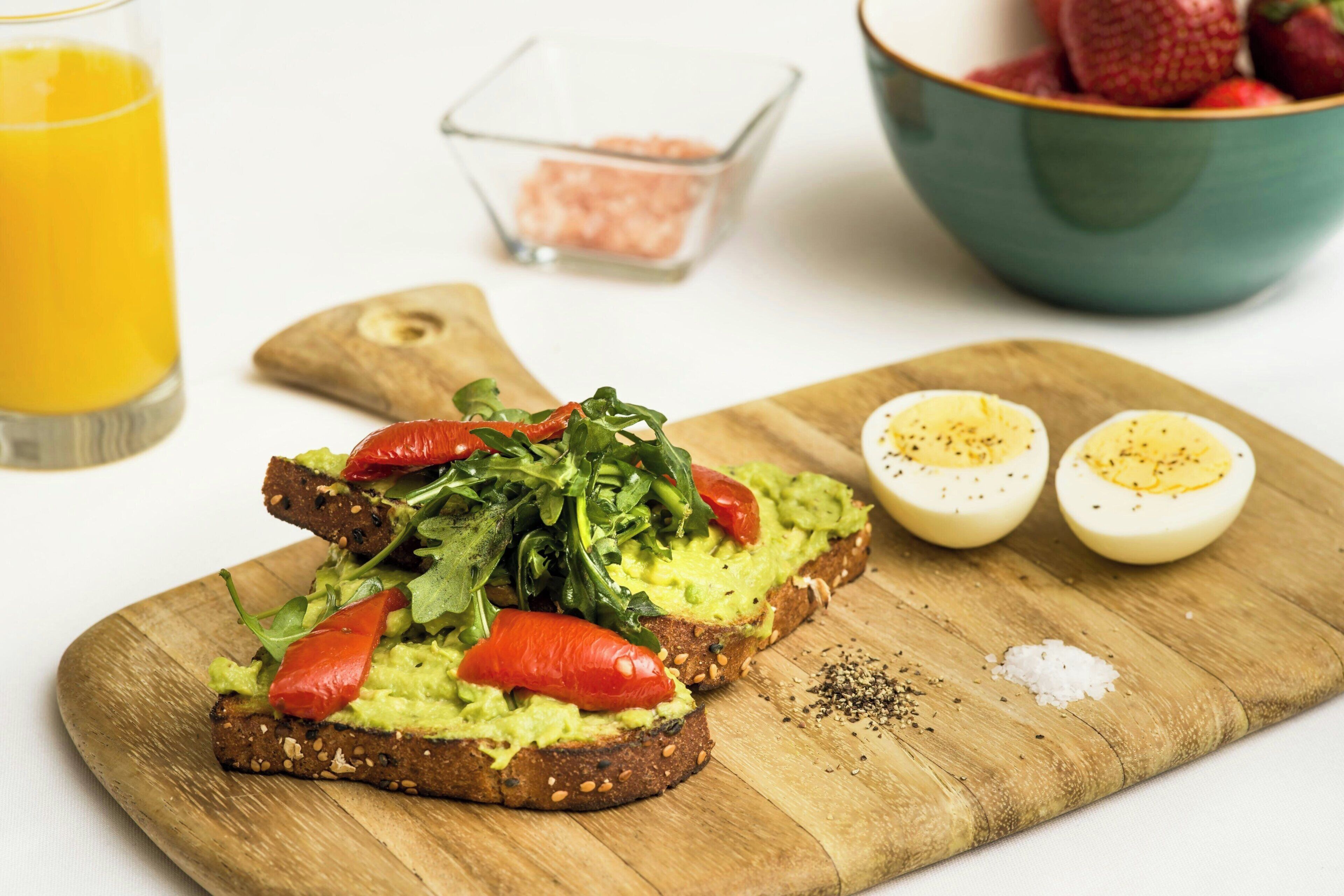 A wooden cutting board with a sandwich topped with avocado, tomato, and greens, a bowl of strawberries, and a glass of orange juice.