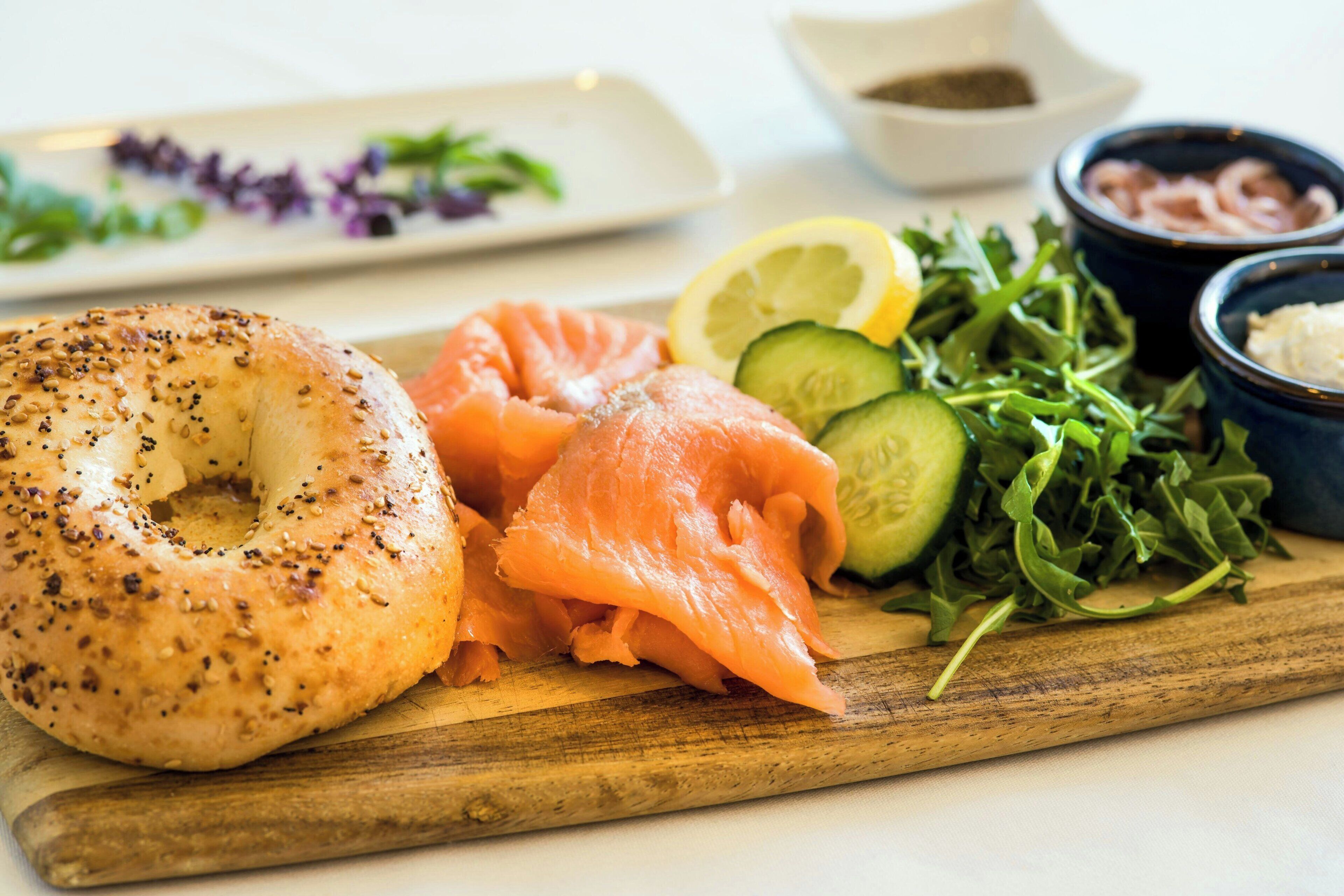 Een maaltijd met een bagel, gerookte zalm, plakjes komkommer, rucola, een citroen en twee kleine schaaltjes met toppings.
