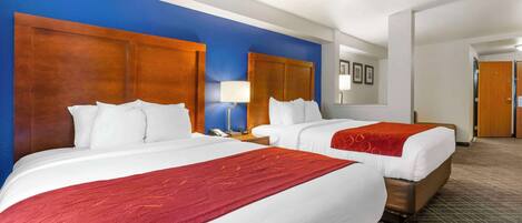 A hotel room with two beds, a wooden headboard, a red throw blanket, a nightstand with a lamp, and a door in the background.