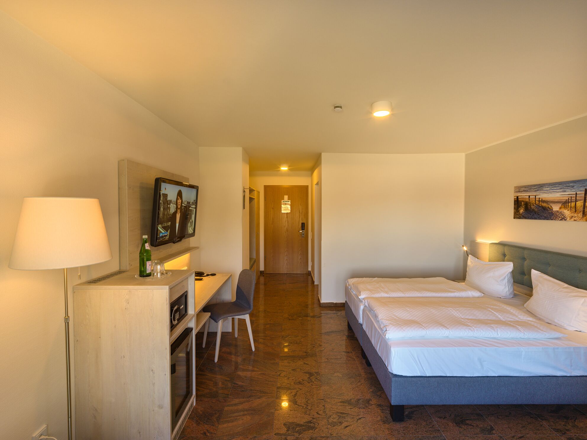 A hotel room with a bed, a desk with a chair, a television, and a painting on the wall.