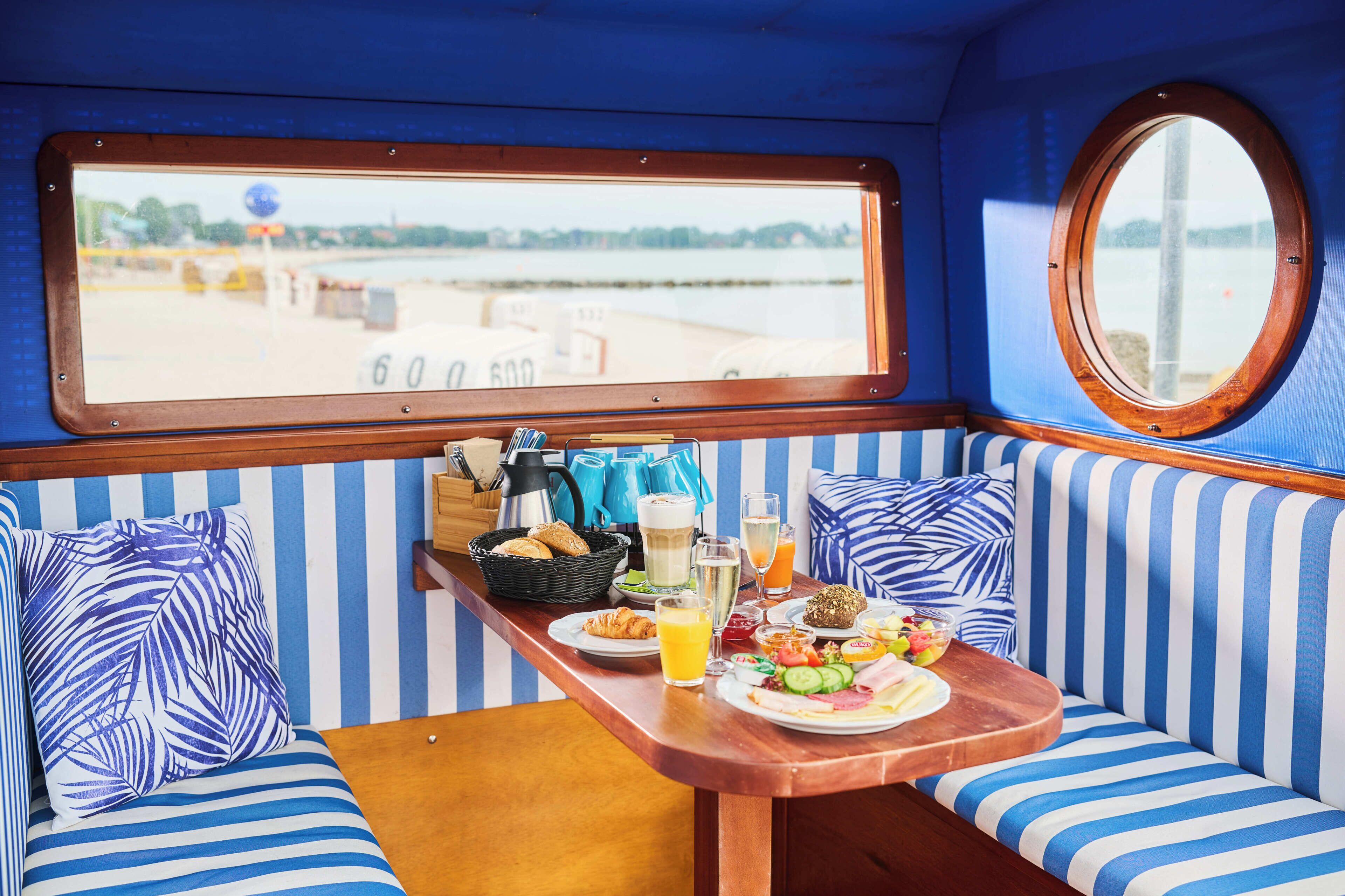 A dining area with striped blue and white walls, a wooden table with breakfast, and a window overlooking a body of water.