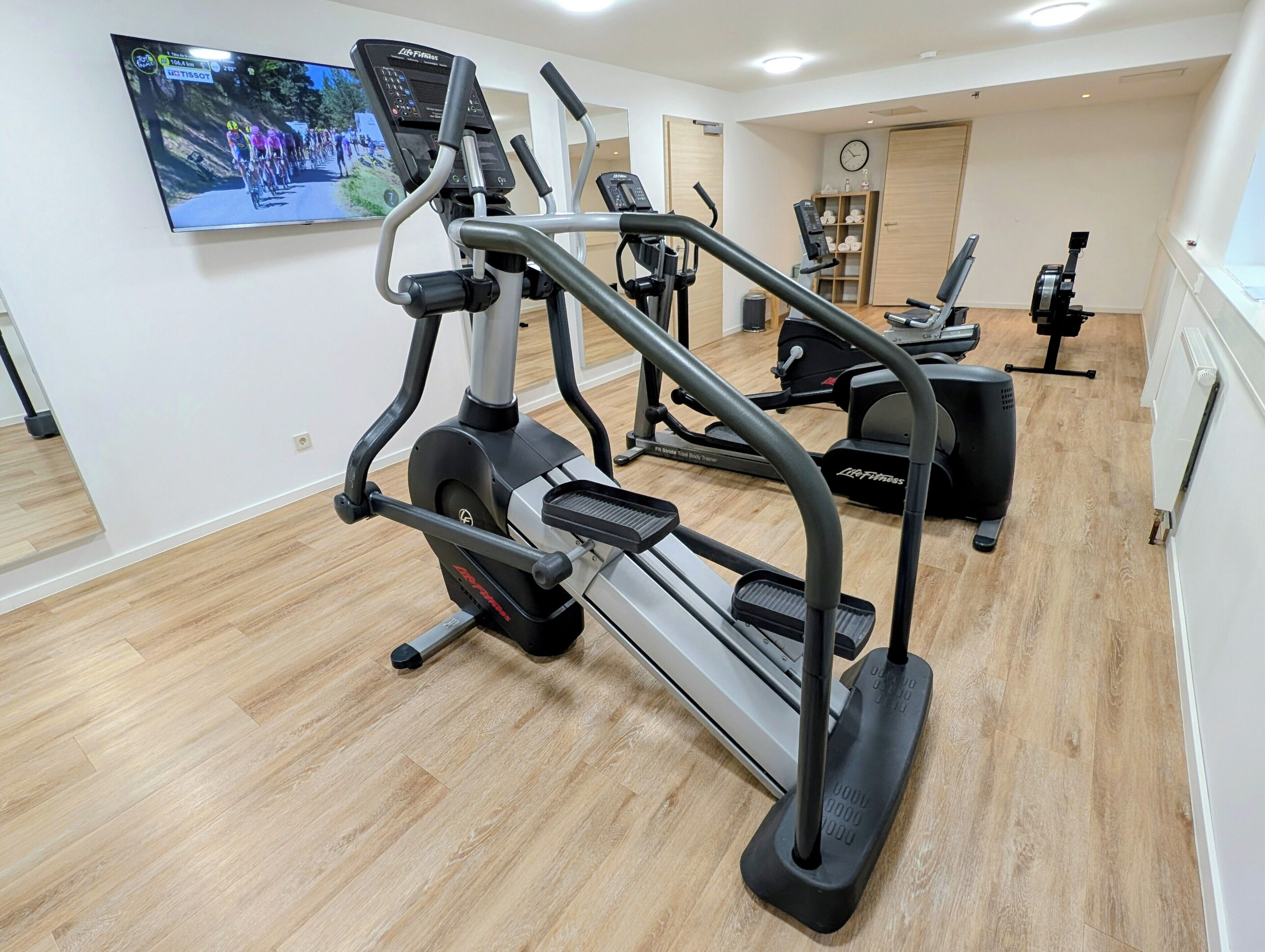 A gym with cardio machines, a TV screen, and a wooden floor.