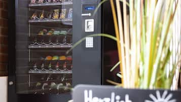 Máquina expendedora con diferentes aperitivos y bebidas, un botón pulsador y un letrero que dice "Herzlich Willkommen".