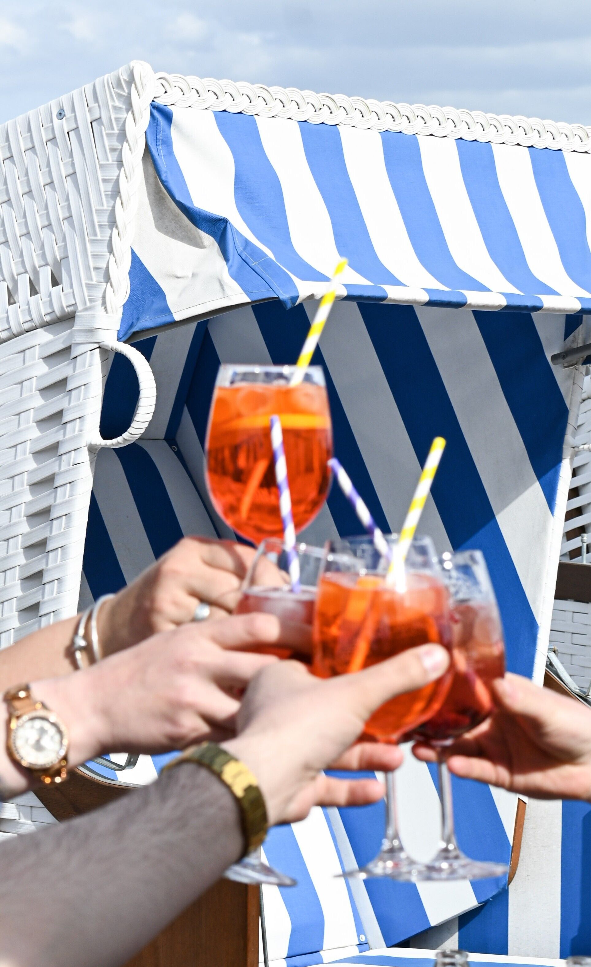 A beach chair with a striped awning, a glass of orange drink, and a watch on a wrist.