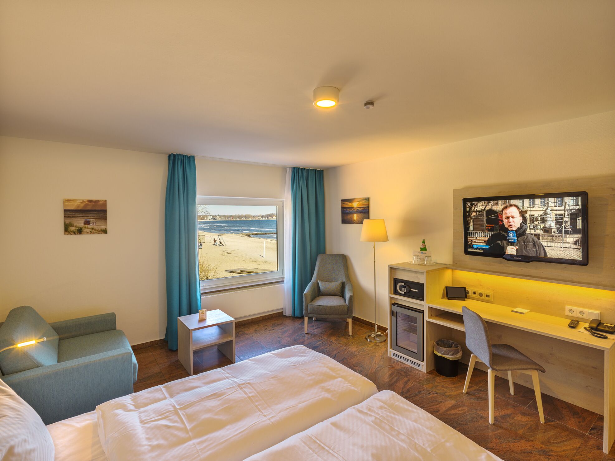 A hotel room with a bed, a desk, a chair, a TV, and a window with a view of the beach.