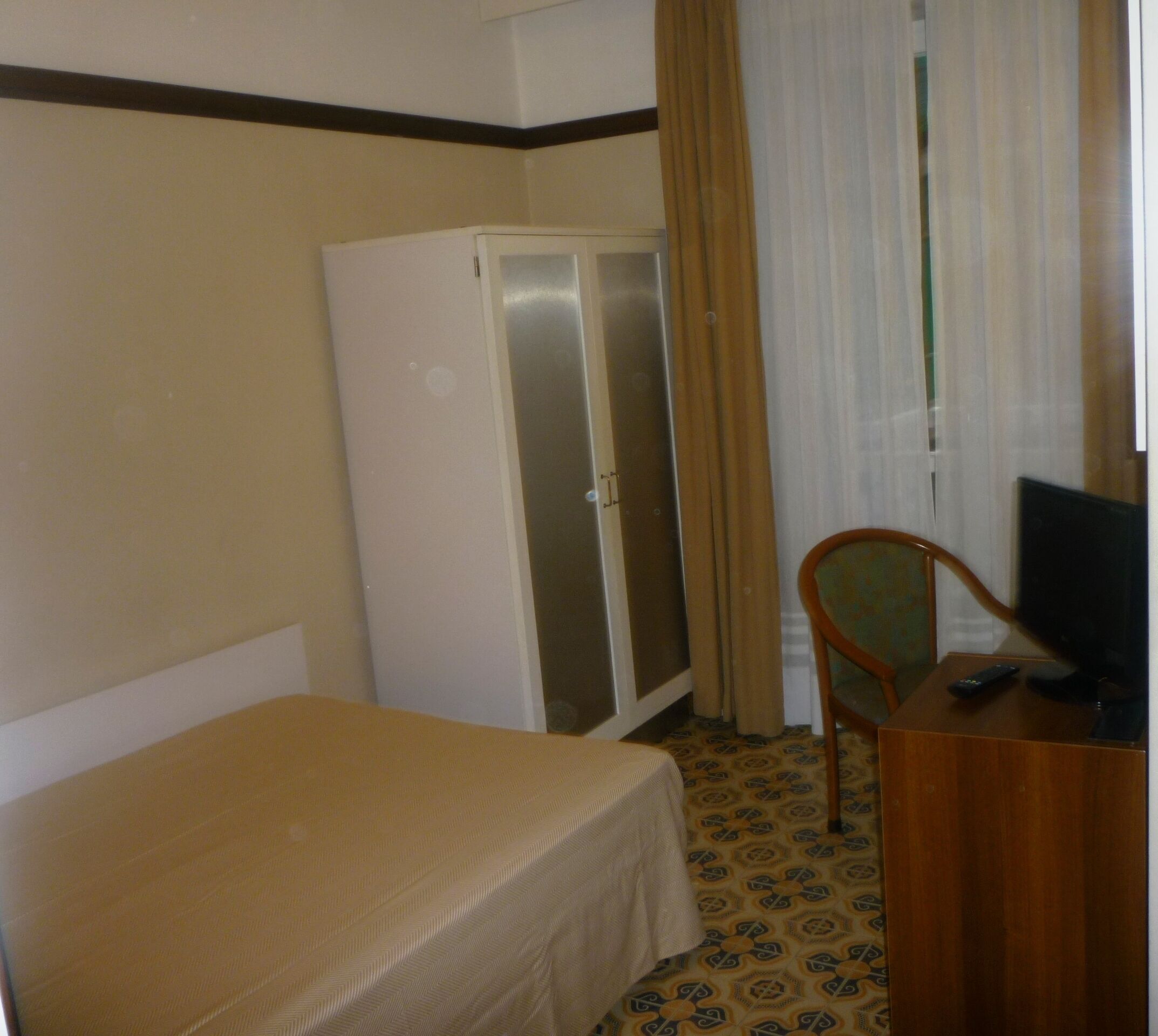 A hotel room with a single bed, a wardrobe, a desk with a television, and a chair.