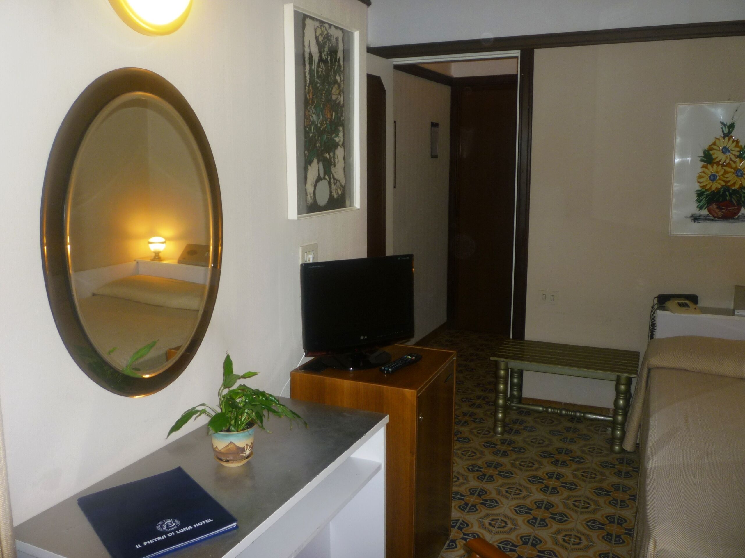 A hotel room with a bed, a desk with a computer, a TV, a mirror, a plant, and a framed picture on the wall.