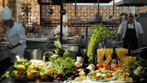 Una cocina con chefs preparando comida, diversas verduras y pasta exhibidas en la encimera.
