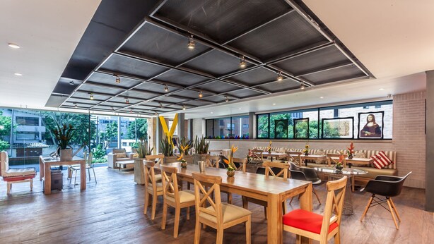 A spacious dining area with wooden tables and chairs, large windows with artwork, and a view of greenery outside. - Mika Suites (Bogotá)