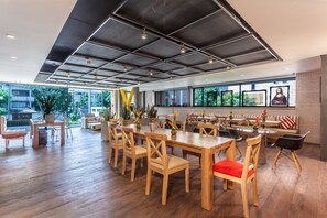 A spacious dining area with wooden tables and chairs, large windows with artwork, and a view of greenery outside. - Mika Suites (Bogotá)
