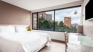 A modern hotel room with a large bed, a desk, and a view of buildings and trees through a large window. - Mika Suites (Bogotá)