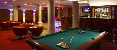 A pool table with a cue stick, a bar area with bottles, and red chairs in a room with columns and a carpeted floor.
