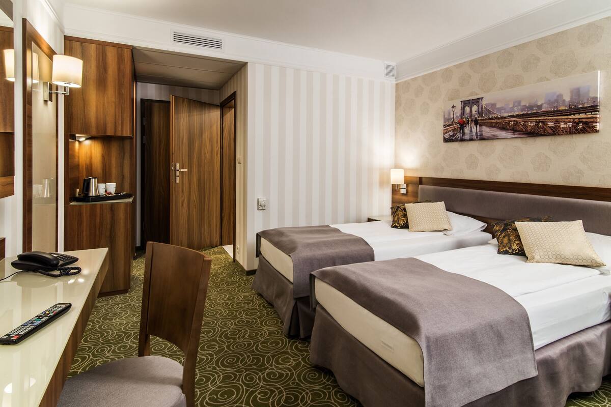 Une chambre d’hôtel avec deux lits, un bureau avec un téléphone, une chaise, un tableau représentant un pont et une moquette à motifs.