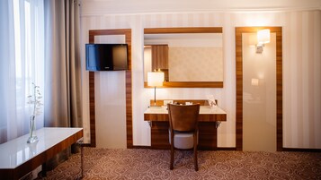 A hotel room with a flat-screen TV, a desk with a chair, a vanity with a mirror, and a patterned carpet.