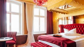 Una habitación de hotel con una cama grande, un sillón tapizado rojo, una mesita pequeña y un ventanal con cortinas.