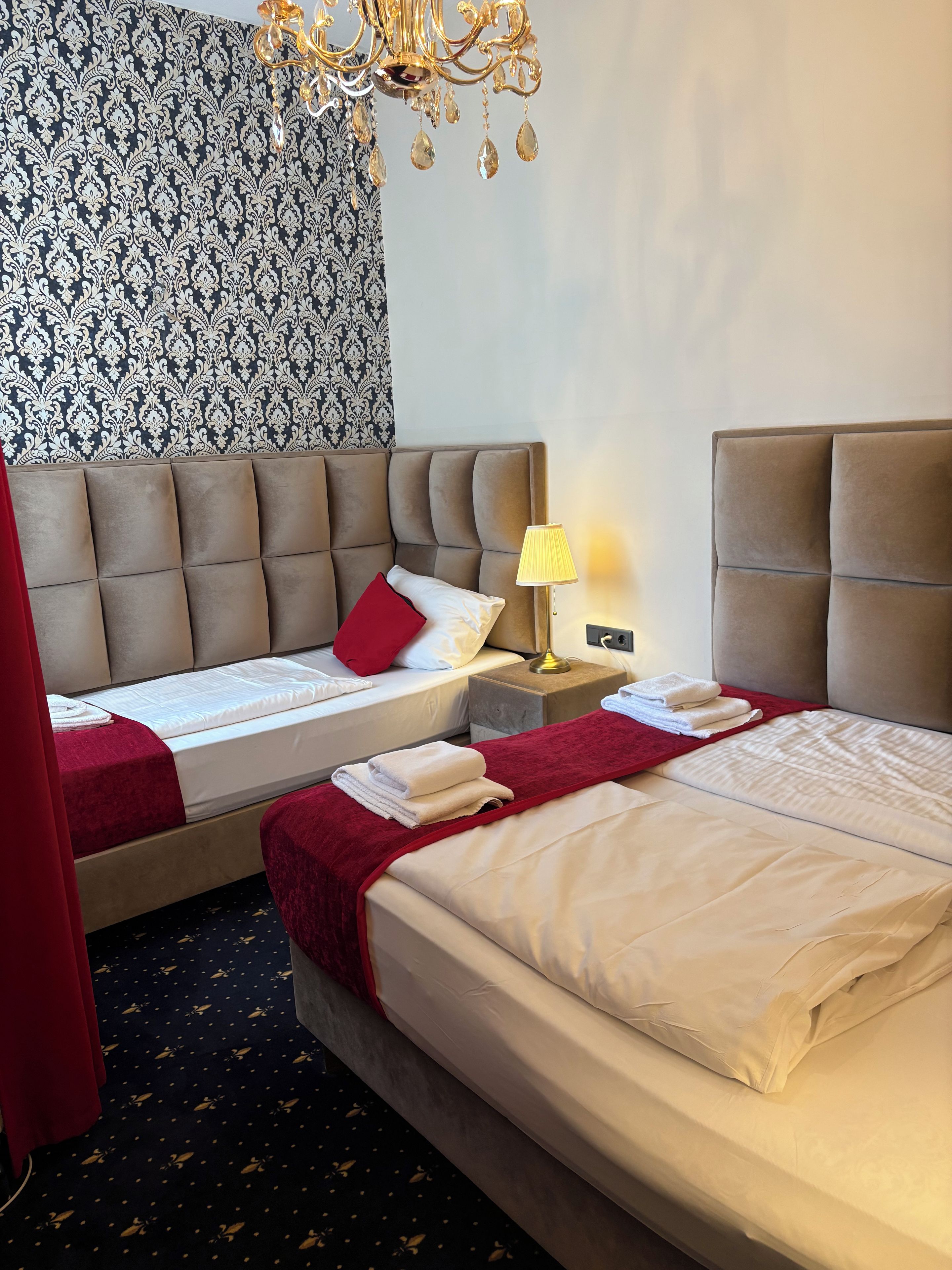 A hotel room with two beds, a chandelier, a patterned wallpaper, and a red curtain.