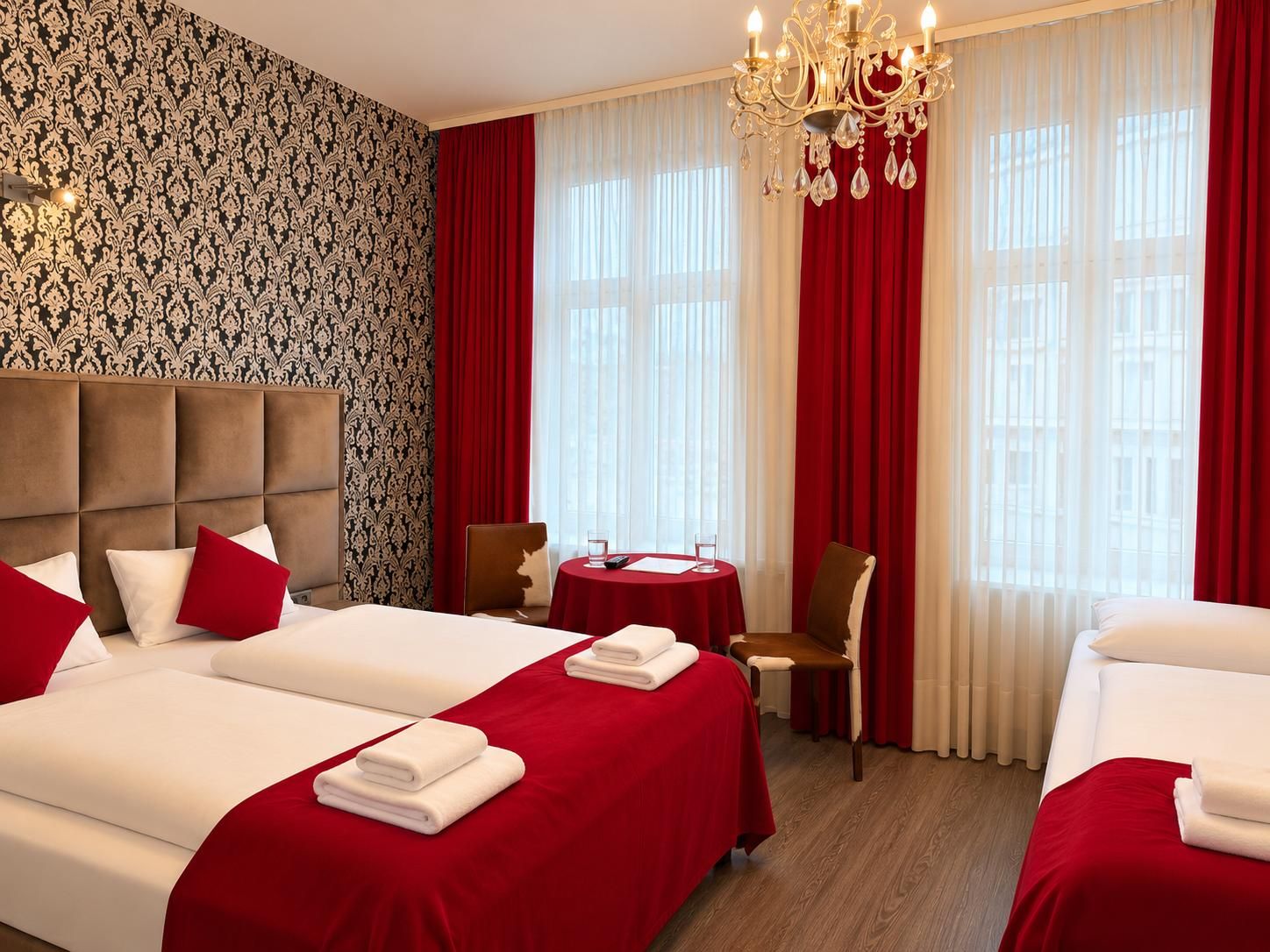 Une chambre d’hôtel avec deux lits, une table à manger, un lustre et une fenêtre avec des rideaux.