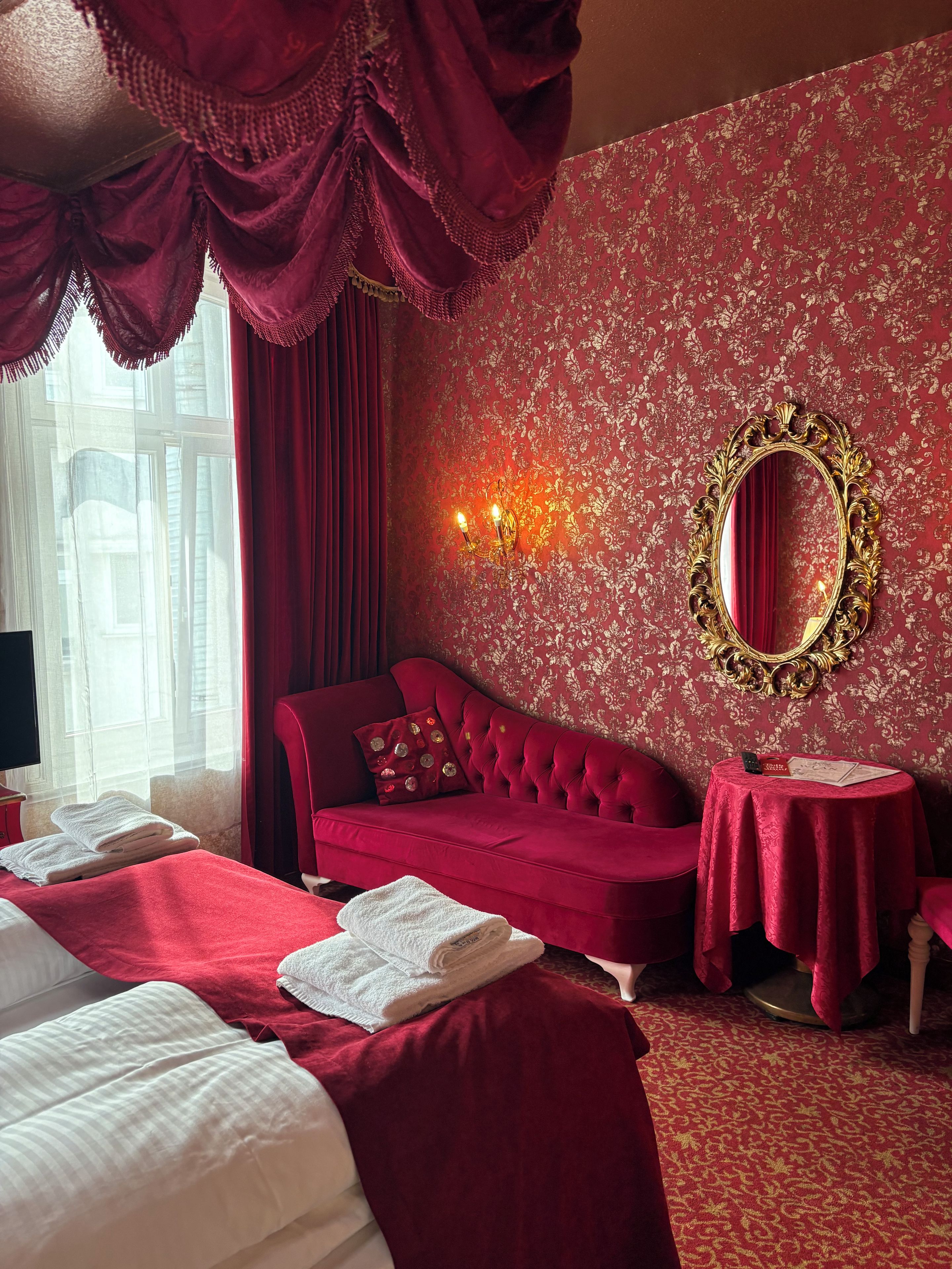 Une pièce avec un canapé rouge, un lit recouvert de draps blancs, une table ronde avec une nappe rouge et un miroir mural encadré d’or.