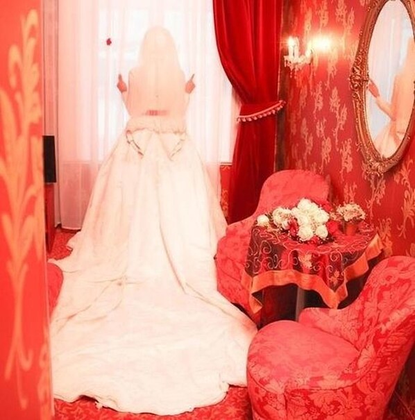 A room with a red carpet, a white dress on a mannequin, a red chair, and a table with flowers. - Hotel Village (Hamburg)