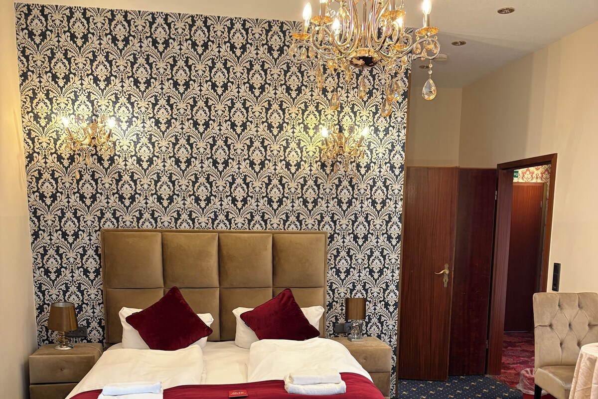 A hotel room with a large bed, a chandelier, patterned wallpaper, and a carpet with a star design.