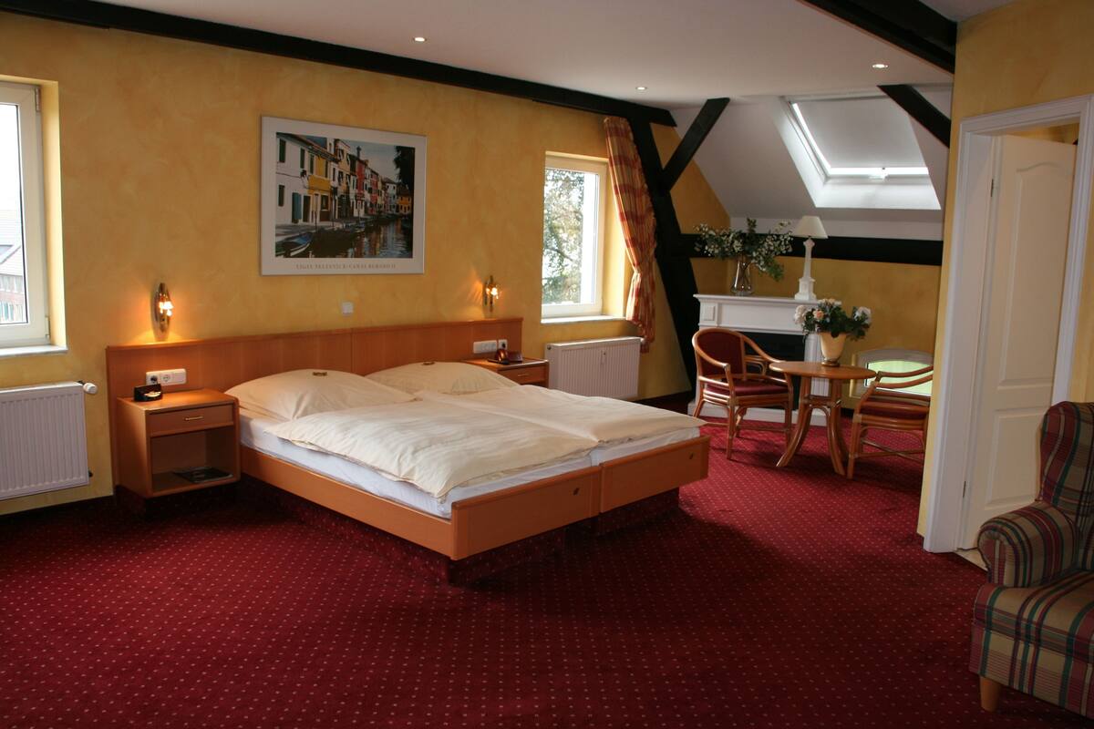 A hotel room with a bed, a small table with chairs, a window with curtains, a radiator, and a painting on the wall.