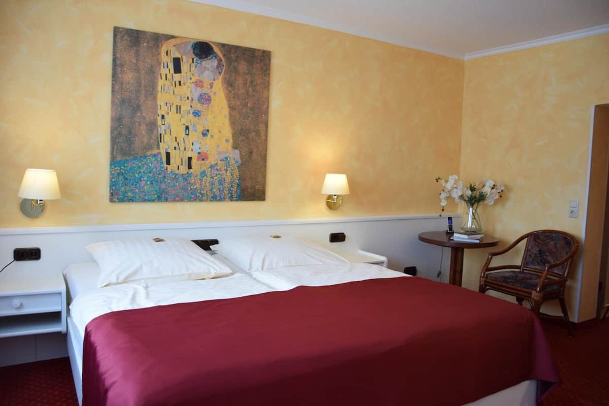 A hotel room with a large bed, two bedside tables, a chair, a painting on the wall, and a vase with flowers.
