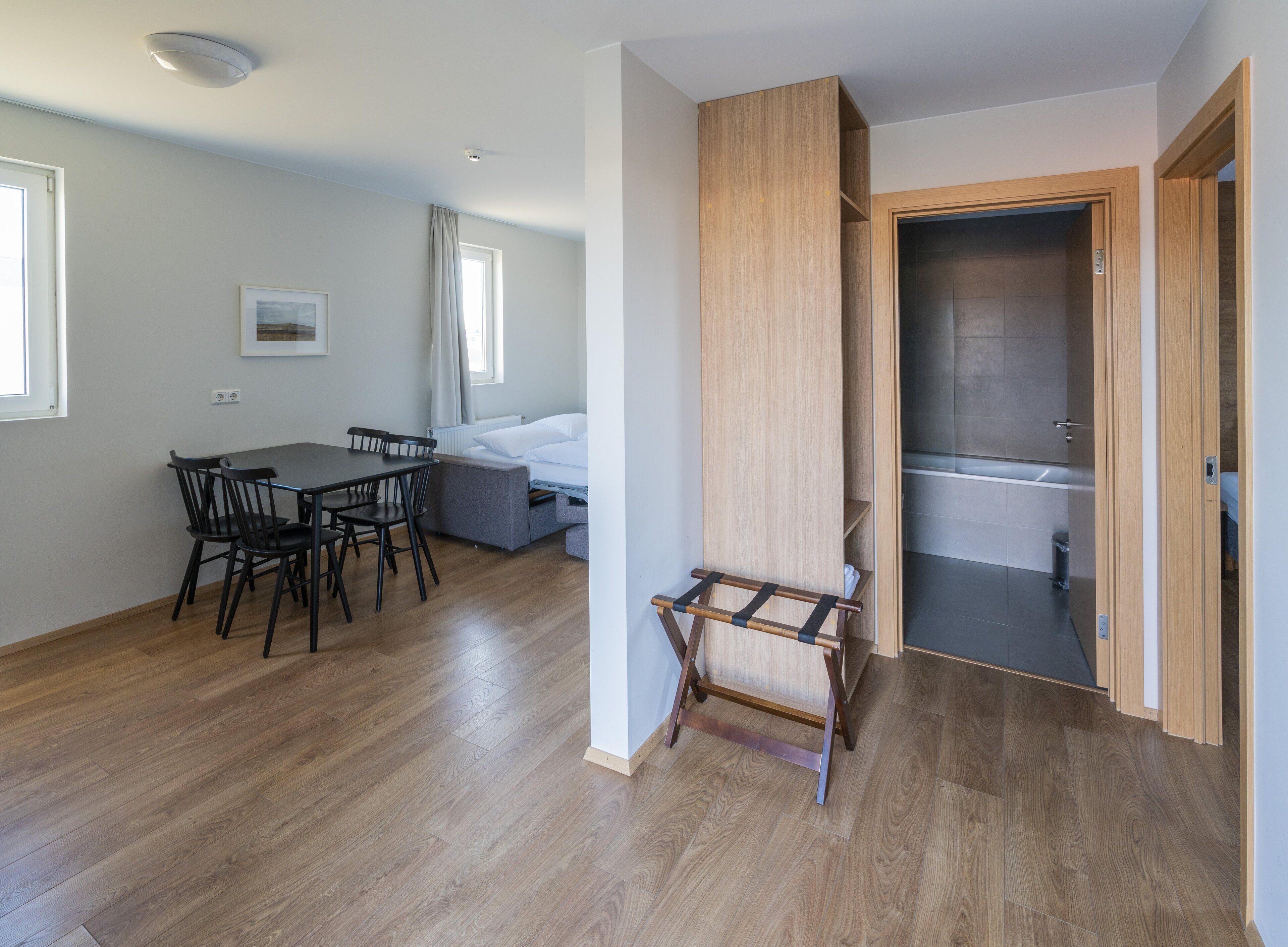 Ein modernes Zimmer mit Holzboden, einem Esstisch mit Stühlen, einer Couch und einem kleinen Holzschemel. Eine Tür führt zu einem Badezimmer mit einer Badewanne.