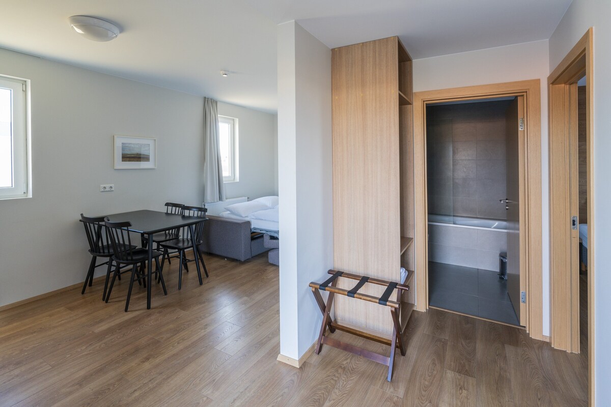 A modern room with a wooden floor, a dining table with chairs, a sofa, and a small wooden stool. There is a door leading to a bathroom with a bathtub.