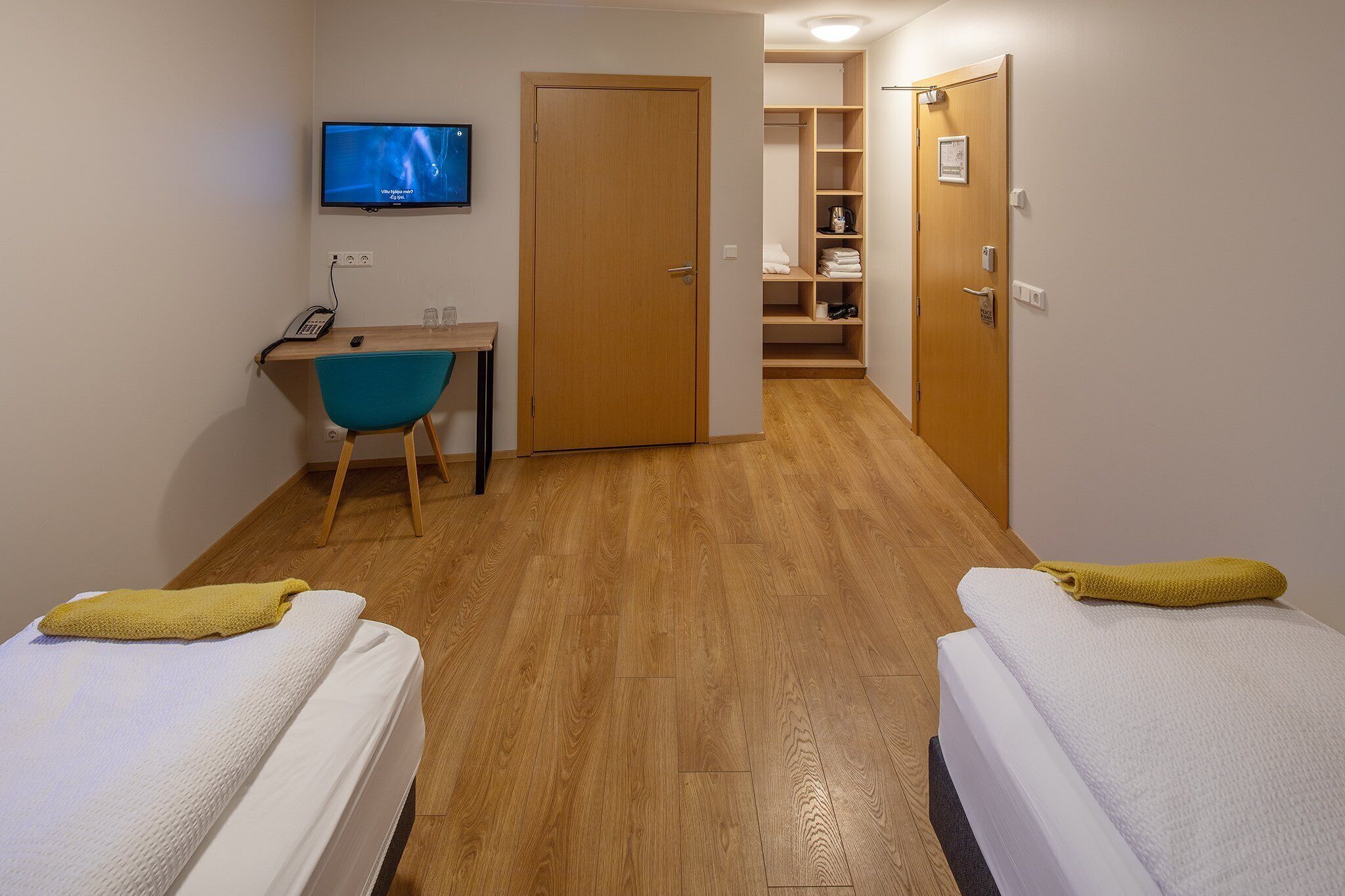 A small, well-lit room with two beds, a desk, a chair, and a television on the wall.