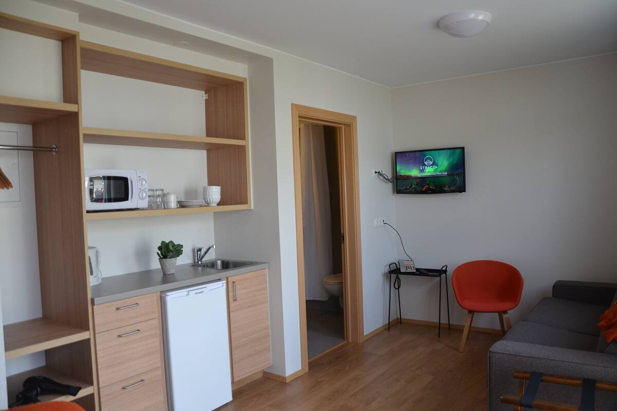 Eine kompakte Küche mit Mikrowelle, Spüle und Schränken, ein kleiner Essbereich mit einem Stuhl und ein an der Wand befestigter Fernseher.