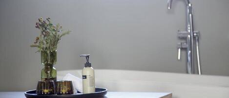 Un meuble-lavabo de salle de bain avec un vase en verre contenant des fleurs, une bouteille de shampoing et deux verres posés sur un plateau.