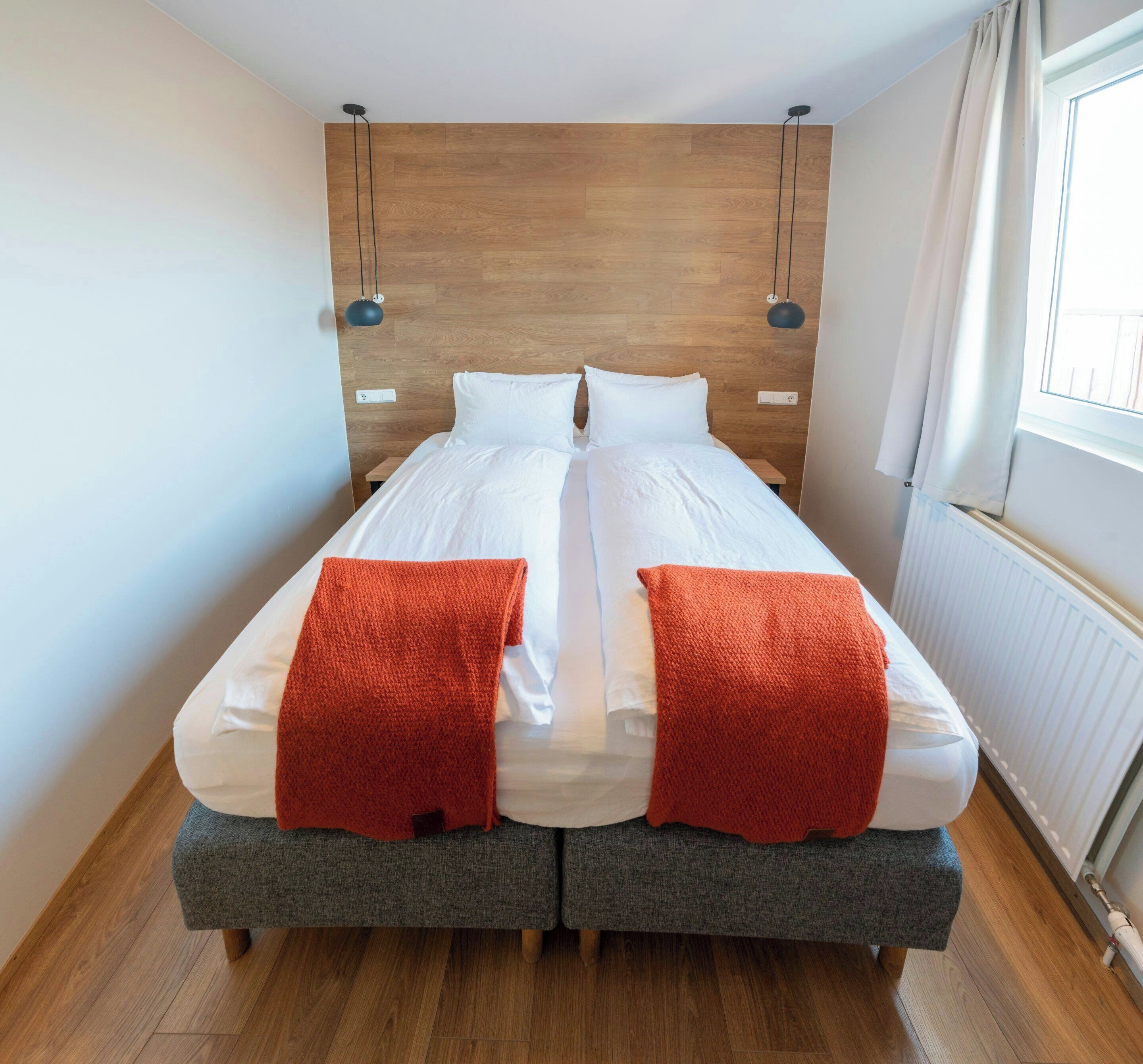 A double bed with a wooden headboard, grey upholstered footboard, and two red throw blankets.