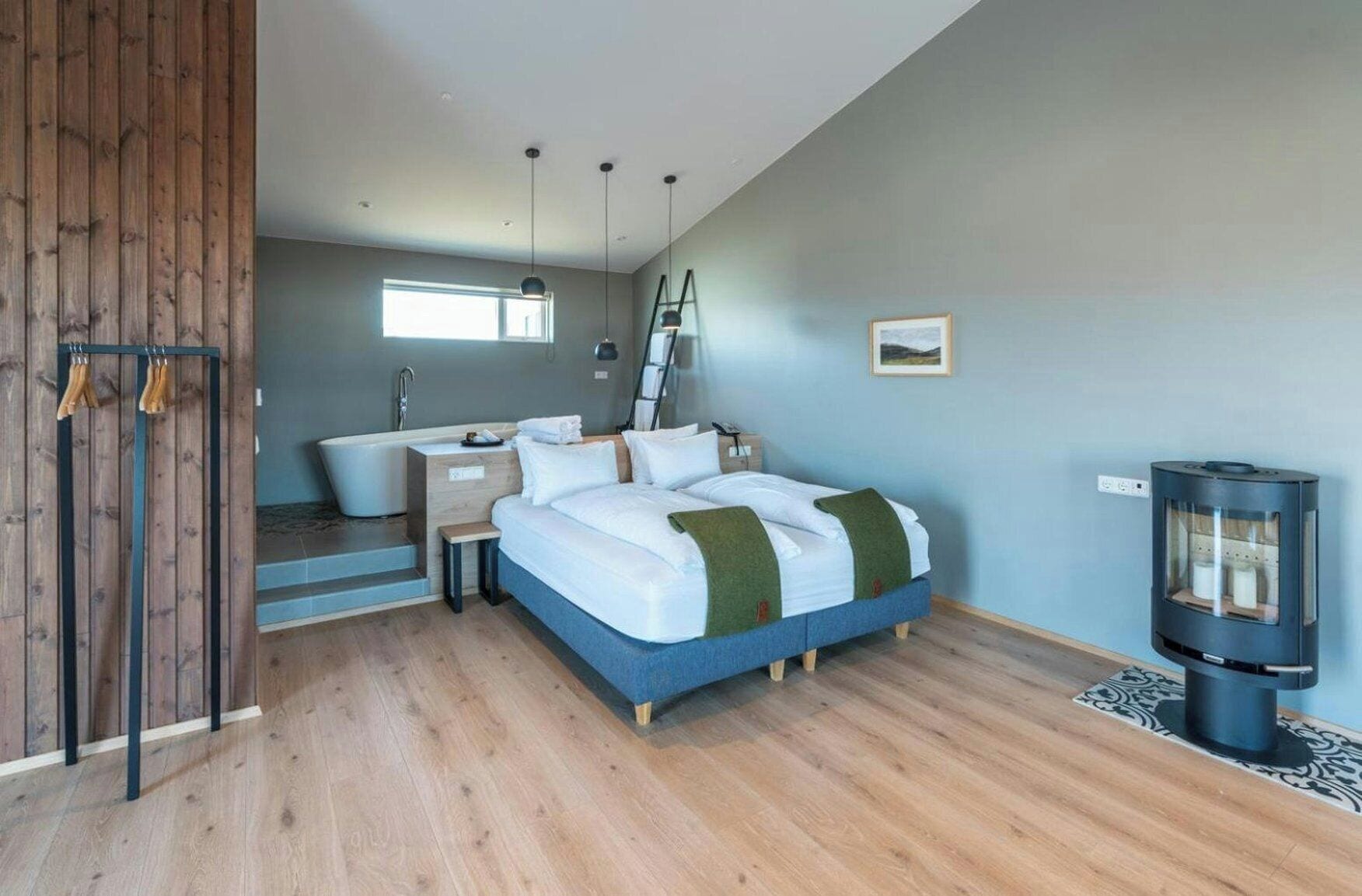 A modern bedroom with a large bed, a wooden floor, a built-in desk, a window, and a wood-burning stove.