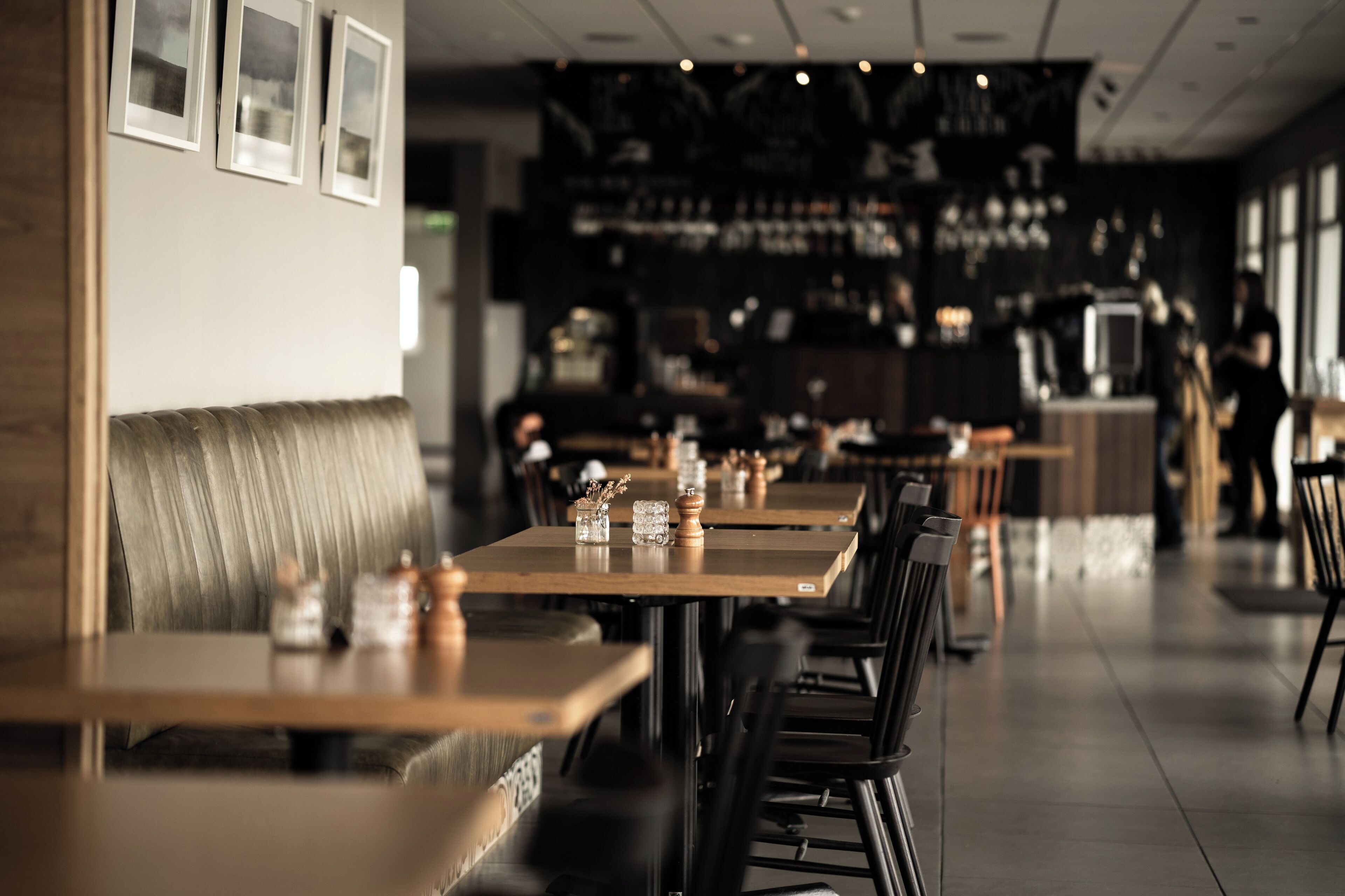 A restaurant with long wooden tables and black chairs, a bar area in the background, and framed pictures on the wall.