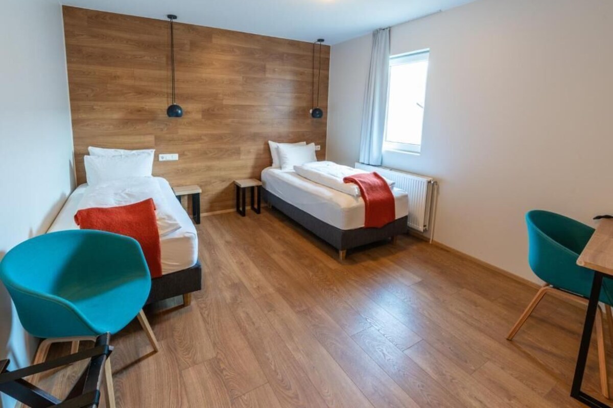 A hotel room with two beds, a wooden headboard, a window with curtains, and a teal chair.