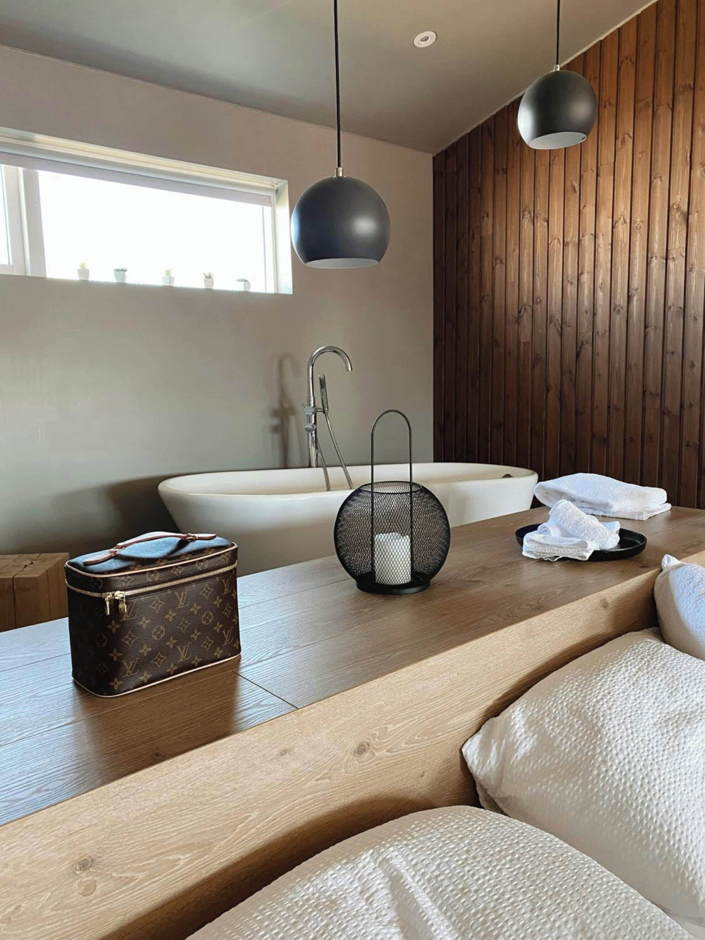 Un baño moderno con una pared de madera, una bañera blanca, una lámpara colgante negra y un bolso Louis Vuitton sobre el mostrador.