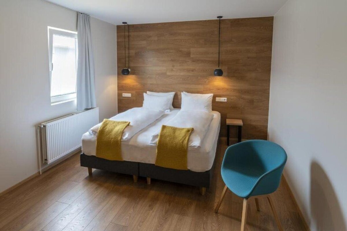 A modern bedroom with a wooden headboard, a double bed with white linens and a yellow throw, a blue chair, and a window with curtains.
