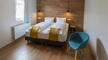A modern bedroom with a wooden headboard, a double bed with white linens and a yellow throw, a blue chair, and a window with curtains.