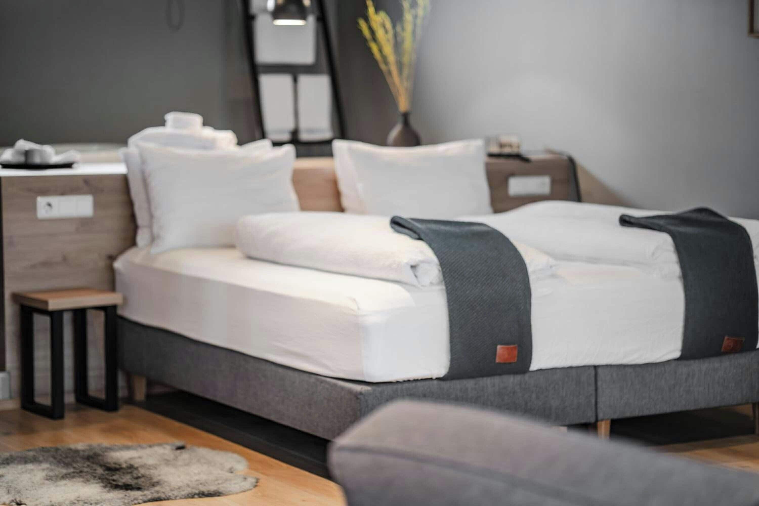 Una cama con estructura gris, ropa de cama blanca y una mesita de noche con lámpara.