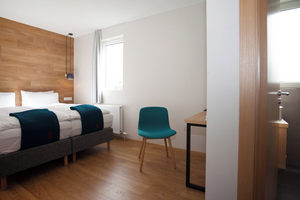 A modern hotel room with a wooden headboard, a bed with white linens and teal throw pillows, a teal chair, a small wooden table, and a window with curtains.