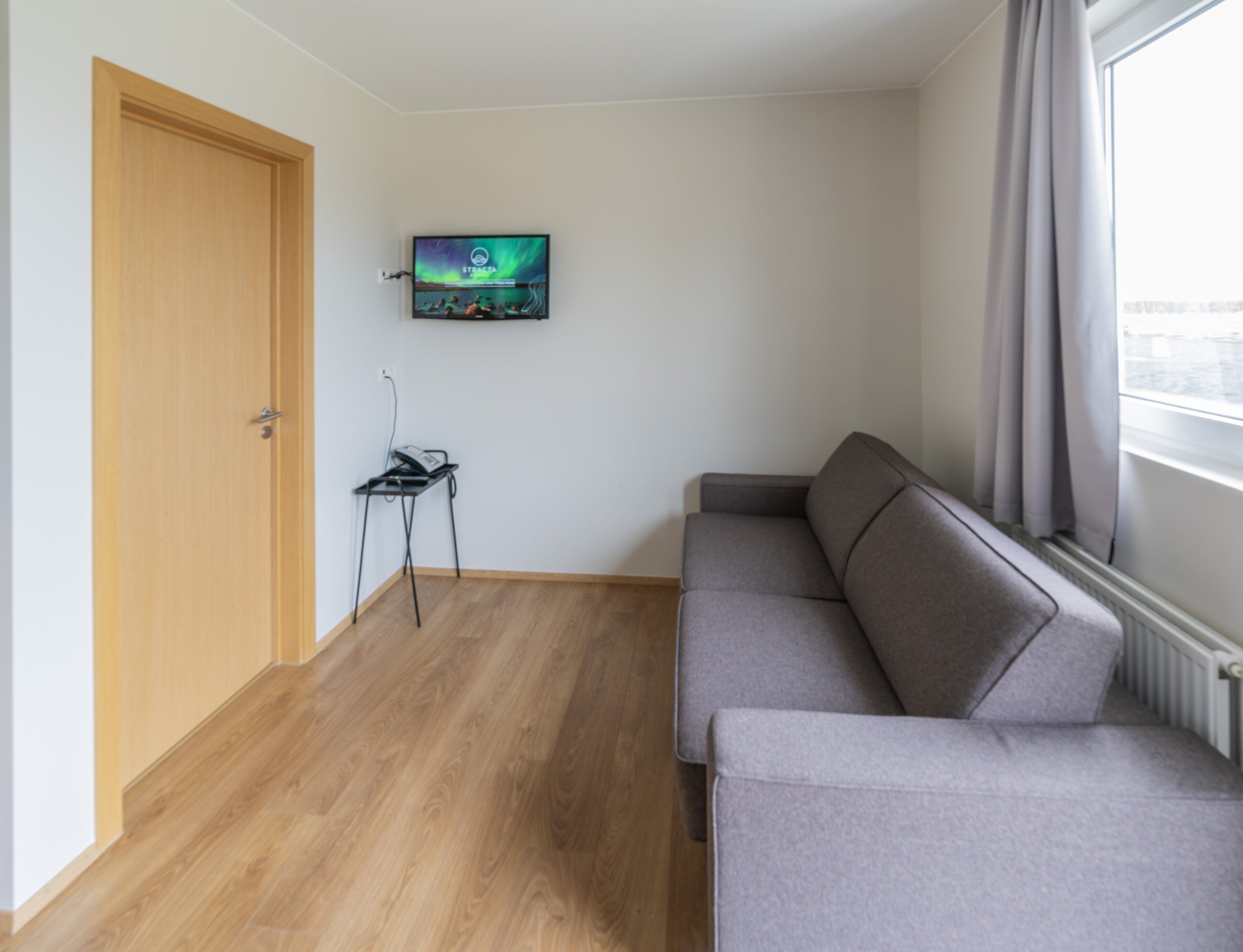 Una sala de estar pequeña y moderna con un sofá gris, una puerta de madera, un televisor montado en la pared y una mesita con un teléfono.