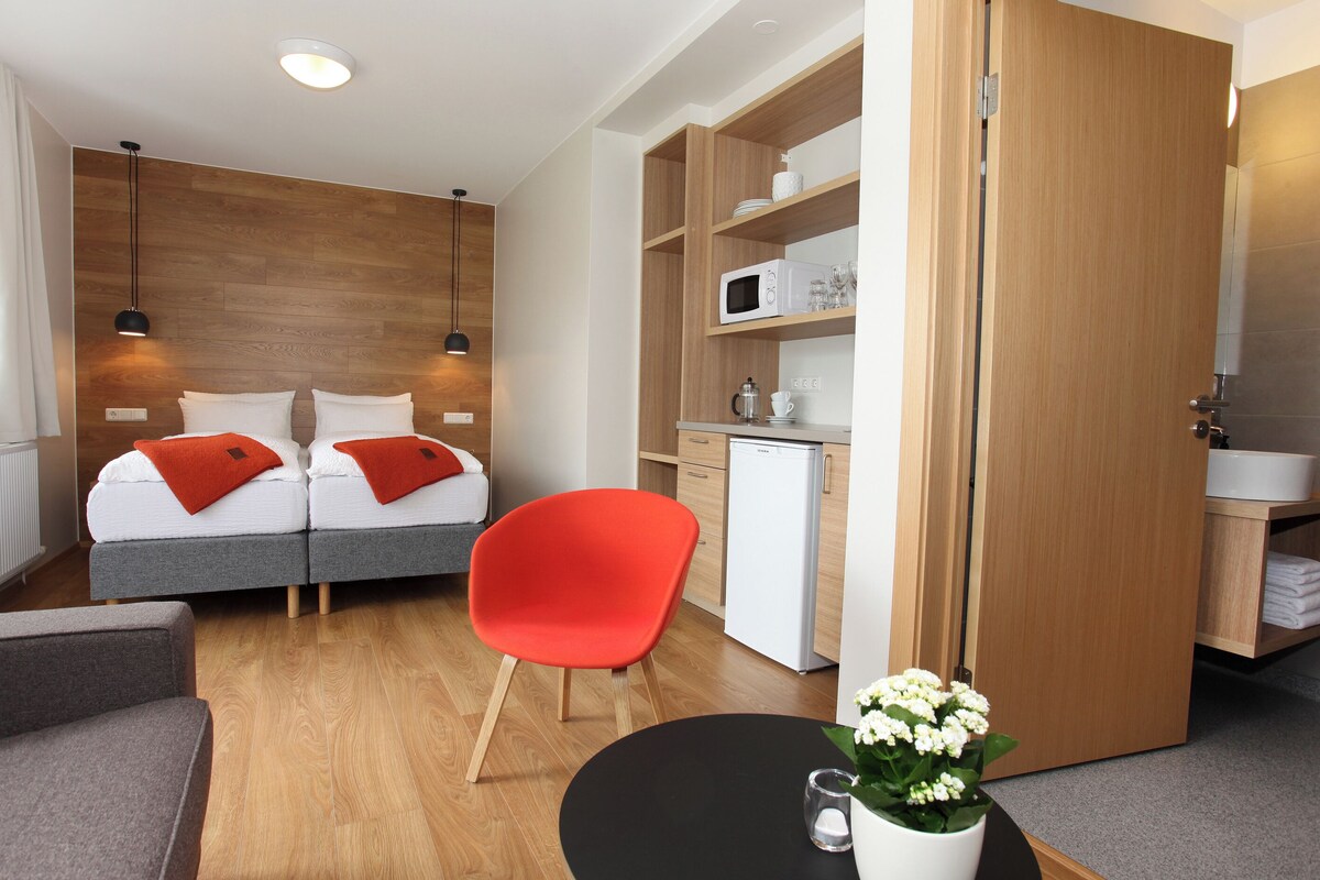 A compact hotel room with a bed, a red chair, a small table with a flower, a microwave, and a compact kitchenette.