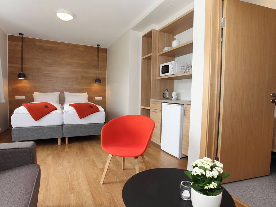 Een kleine hotelkamer met een bed, een rode stoel, een tafeltje met een bloem, een magnetron en een compacte kitchenette.