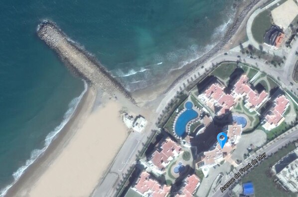 An aerial view of a coastal resort with a beach, a long pier, and a central building complex with a circular pool. - Grand Mogador SEA VIEW (Tangier)