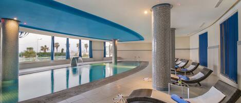 A modern indoor swimming pool with lounge chairs and a view of palm trees.