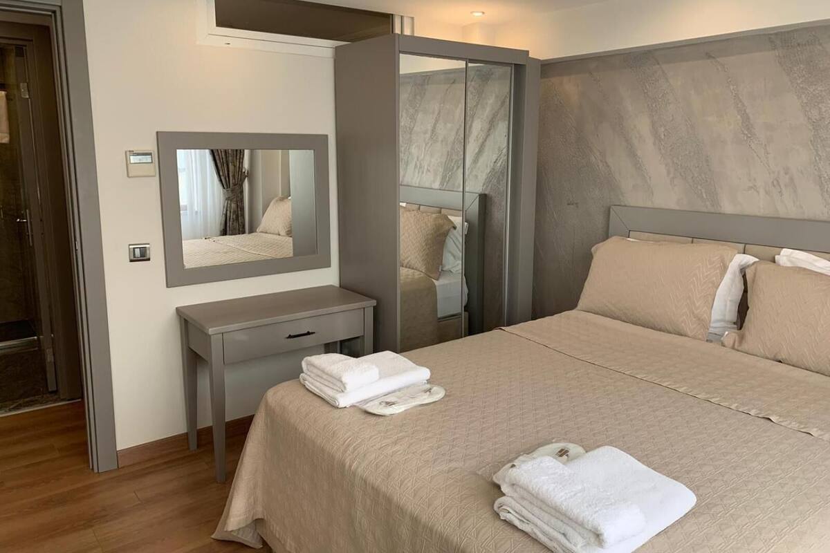 Habitación de hotel con cama, mesita de noche con espejo, armario y baño visible a través de una puerta de cristal.
