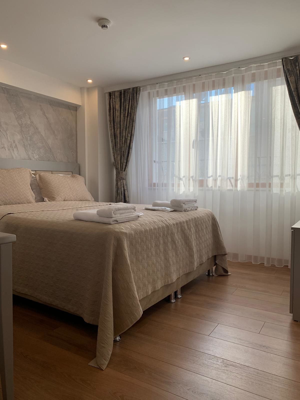 A neatly made bed with a beige bedspread and pillows, a bedside table with a lamp, and a window with sheer curtains.