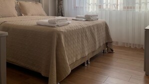 A neatly made bed with a beige bedspread and pillows, a bedside table with a lamp, and a window with sheer curtains. - Regency World Suite Istanbul (Istanbul)