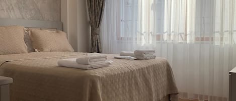 Una cama bien hecha con una colcha color beige y almohadas, una mesita de noche con una lámpara y una ventana con cortinas translúcidas.